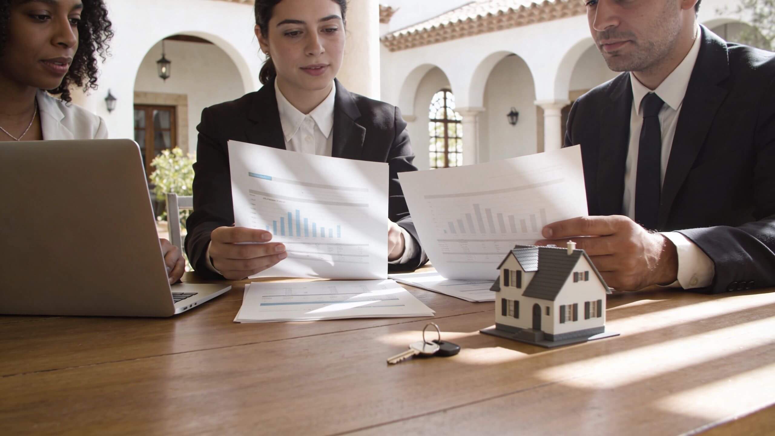 Certificados Energéticos | Aprende que es un Broker Inmobiliario y sus ventajas en 2026 Tres profesionales de negocios revisando informes financieros con un modelo de casa y llaves sobre la mesa.